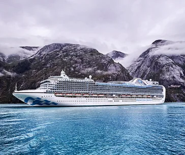 Cruise ship "Ruby Princess" sailing in front of misty Alaskan mountains, with calm blue water in the foreground.