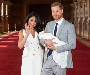 A couple stands inside a grand hall with a red carpet, holding a newborn wrapped in a white blanket.
