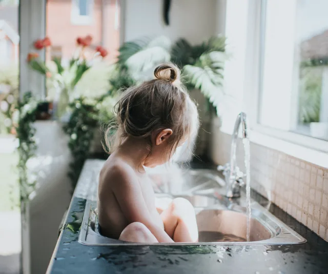 Toddler sitting in a kitchen sink during bath time, surrounded by plants and natural light.