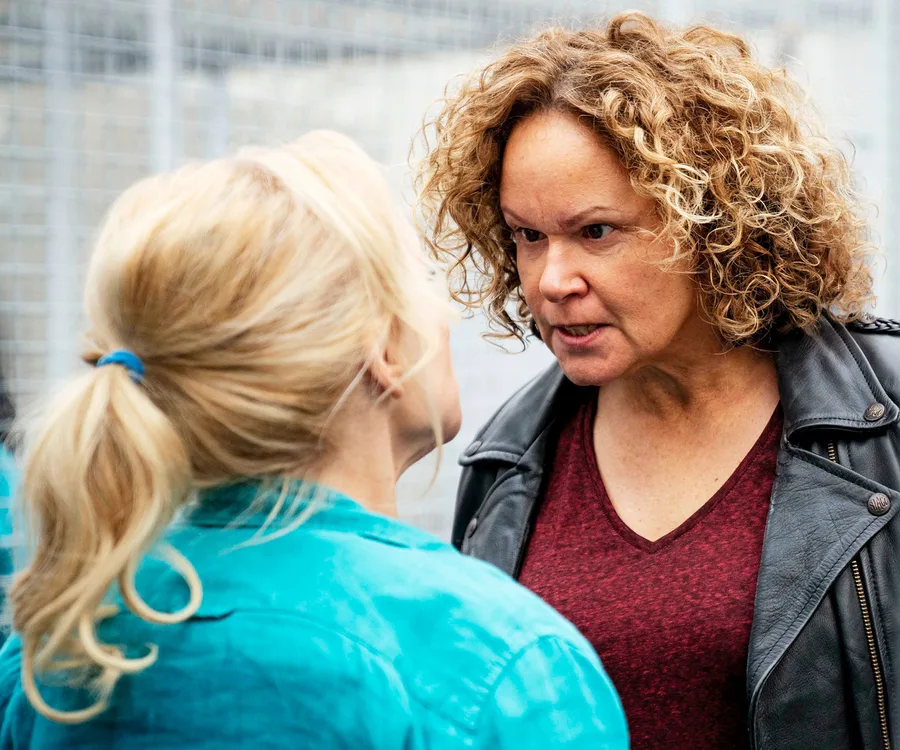 Two women facing each other in a tense exchange. One wears a leather jacket, and the other has a blonde ponytail.