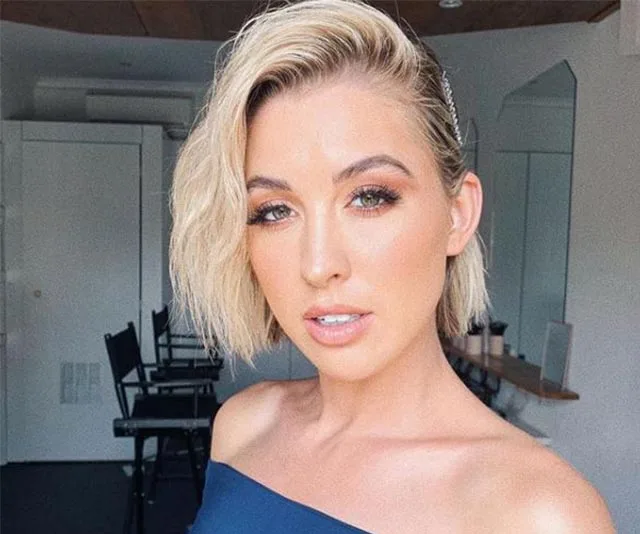 Young woman with short blonde hair and an off-shoulder top, posing in a bright room with mirrors and chairs behind her.