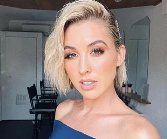 Young woman with short blonde hair and an off-shoulder top, posing in a bright room with mirrors and chairs behind her.