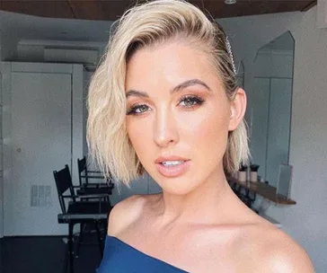 Young woman with short blonde hair and an off-shoulder top, posing in a bright room with mirrors and chairs behind her.
