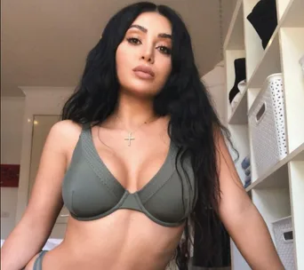 A woman with long dark hair wearing a gray bra poses in a room with shelves and baskets in the background.