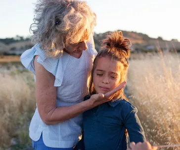 Grandmother comforting upset grandchild in a sunlit field, gently holding their face.