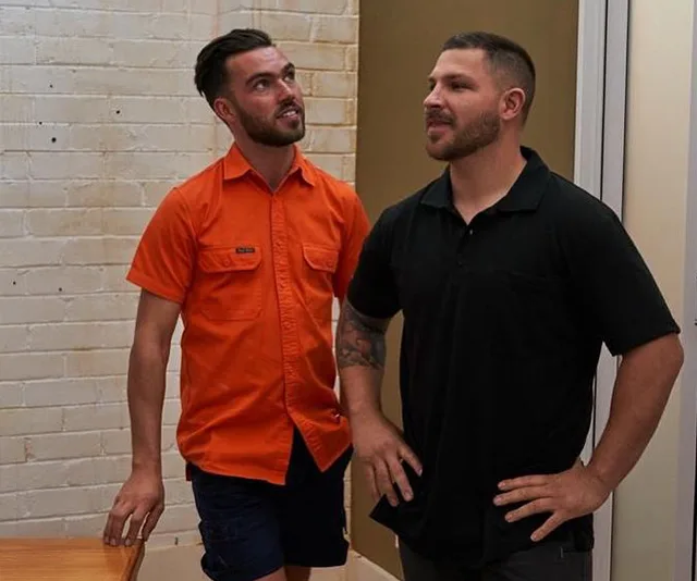 Two men standing indoors, one in an orange shirt and the other in black, conversing and looking to the side.