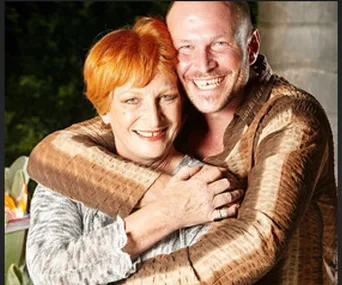 Elderly woman with short red hair hugging a smiling man in a garden, both wearing patterned shirts.