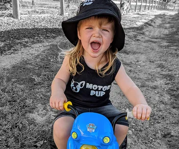Toddler with a mullet wearing a "Motor Pup" shirt and bucket hat, excitedly riding a blue toy motorcycle outdoors.