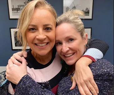 Two women smiling and hugging in a warmly lit room with framed pictures on the wall behind them.