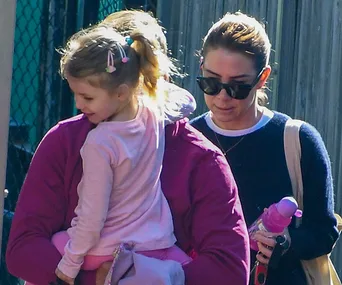 Two women and a young girl with a ponytail and pink outfit. One woman holds the child while the other walks next to them.