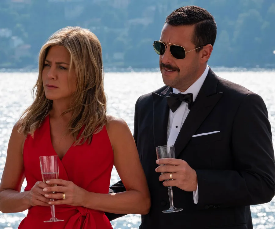 Two people in formal attire holding champagne glasses, standing by a body of water on a sunny day.