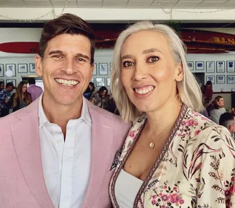 Man in pink blazer and woman in floral top smiling at a casual event with framed photos in the background.