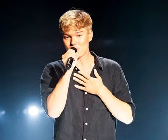 A person singing on stage with a microphone, wearing a black shirt against a dark background with bright lights.