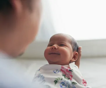 A baby wrapped in a floral blanket smiles while being held by an adult, showing an early connection.