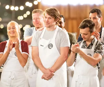 Contestants wearing aprons clap on a cooking show set, smiling under string lights.