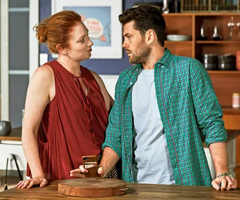 A woman in a red top talks to a man in a green plaid shirt, holding a phone, in a kitchen setting.