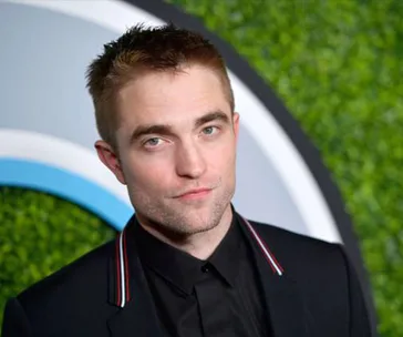 Person in a black suit with short hair stands in front of a green backdrop, looking towards the camera.