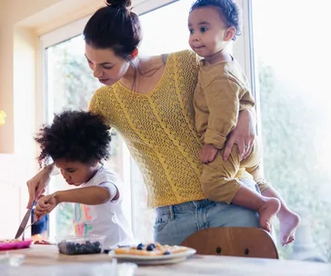 A mother holds her child and helps another child cut food at a table, with sunlight streaming through a window.