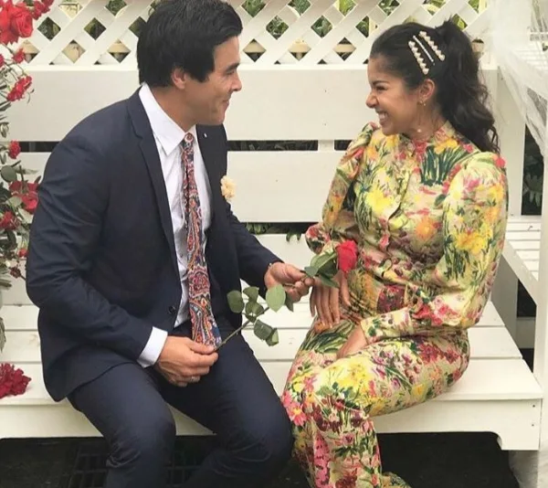 Man in suit gives a rose to a smiling woman in a colorful dress, sitting on a white bench with a floral backdrop.