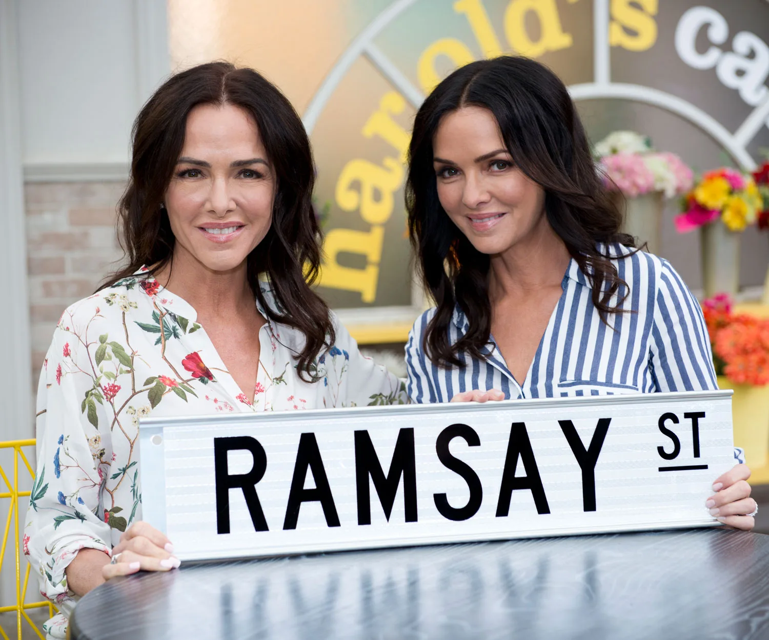 Two women holding a "Ramsay St" sign with flowers in the background.