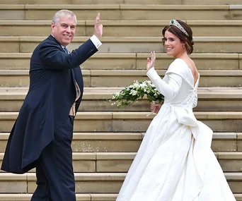 Bride in white gown and man in suit wave while ascending stairs, holding a bouquet.