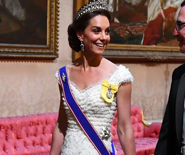 Elegant woman in a white gown and tiara at a formal event, smiling in an ornate room with paintings.