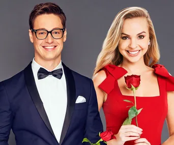 Two smiling individuals in formal attire holding red roses, with a gray background.