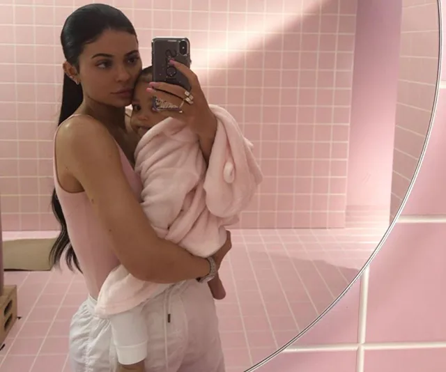 Woman taking a mirror selfie holding a baby wrapped in a pink blanket, against a pink tiled background.