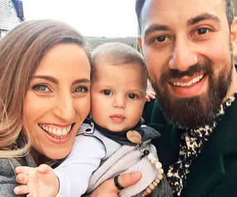 A smiling couple holding a baby, posing for a selfie outdoors with a house in the background.