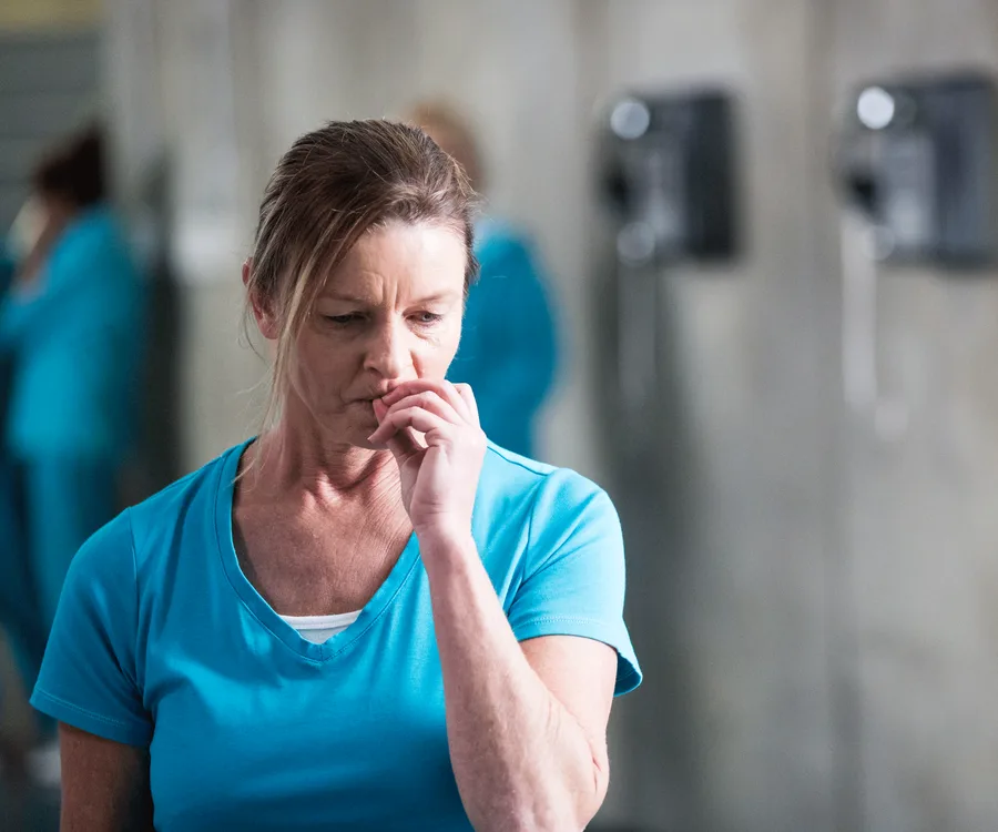 Female character in blue shirt, looking pensive and biting her nail in a dimly lit room with blurred background.