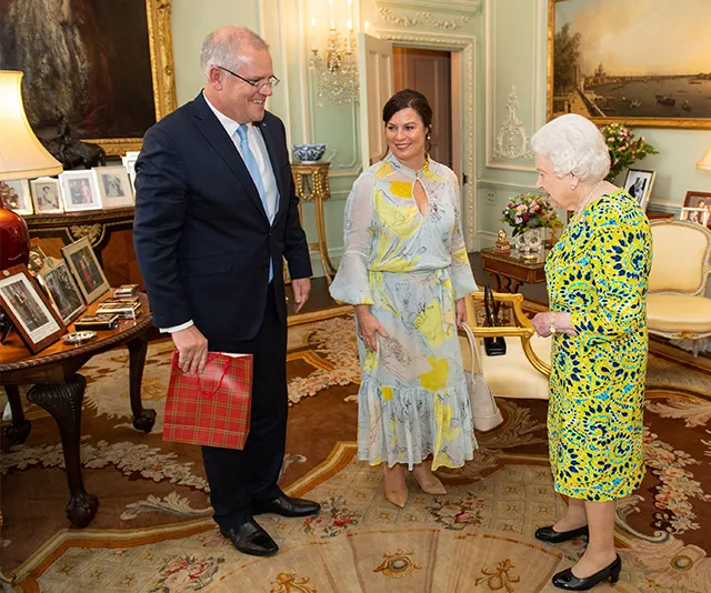 Meeting between three people in an ornate room, with one holding a gift and another in a floral dress.