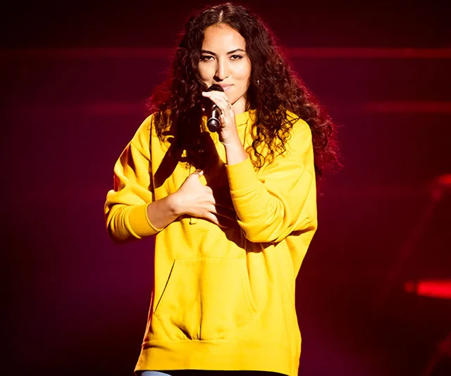 A person with curly hair in a yellow hoodie singing into a microphone on stage with a red background.