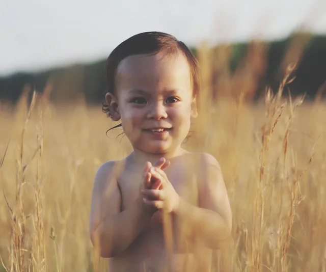 A smiling toddler standing in a sunlit field of tall grass.