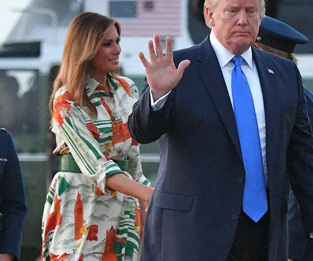 A woman in a colorful dress smiles as she walks with a man in a suit, who raises his hand, near a helicopter.