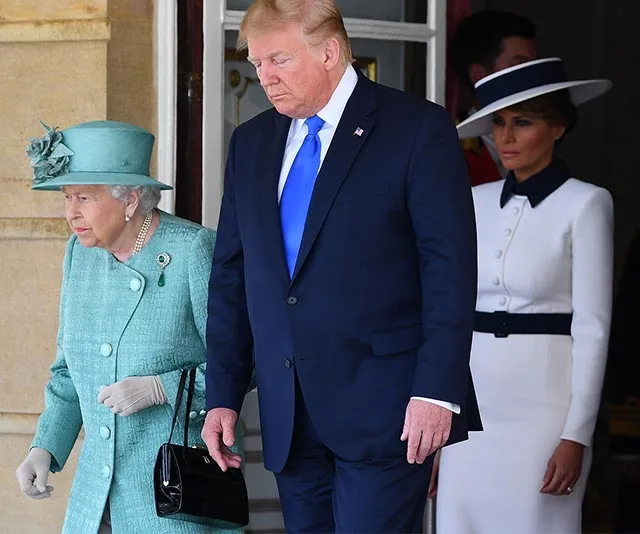 Queen Elizabeth in teal outfit and hat with handbag, walking with two others in formal attire.