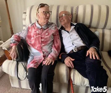Two elderly people sitting on a couch, smiling at each other, in a cozy room.