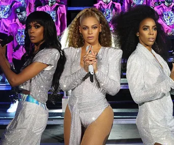 Three women in silver outfits perform on stage, each holding microphones, with backup singers in pink behind them.