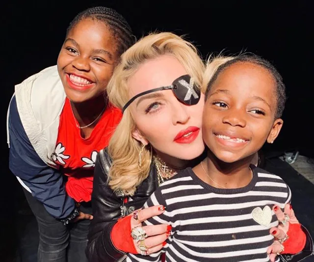 Madonna wearing an eyepatch poses happily with two smiling children, one in red and one in striped top, against a dark background.