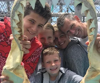 A family of five smiling through a shark jaw frame, outdoors with a metal fence in the background.