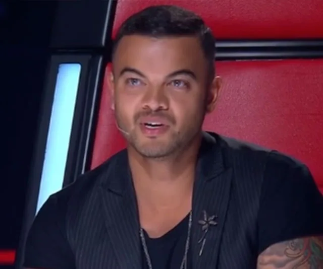 A person with short hair and a dark shirt sits in a red chair, appearing to speak or judge on a TV show.