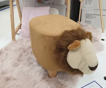 Lion-shaped ottoman with plush tan fabric and a mane, placed on a pale pink rug, near cushions and a wooden frame.