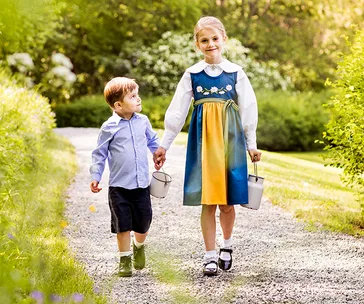 Princess Estelle and Prince Oscar of Sweden walk on a garden path, carrying small buckets, under dappled sunlight.
