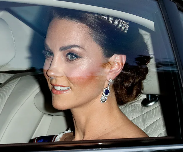 A woman with brown hair, wearing a tiara and sapphire earrings, sits in a car, smiling and looking out the window.