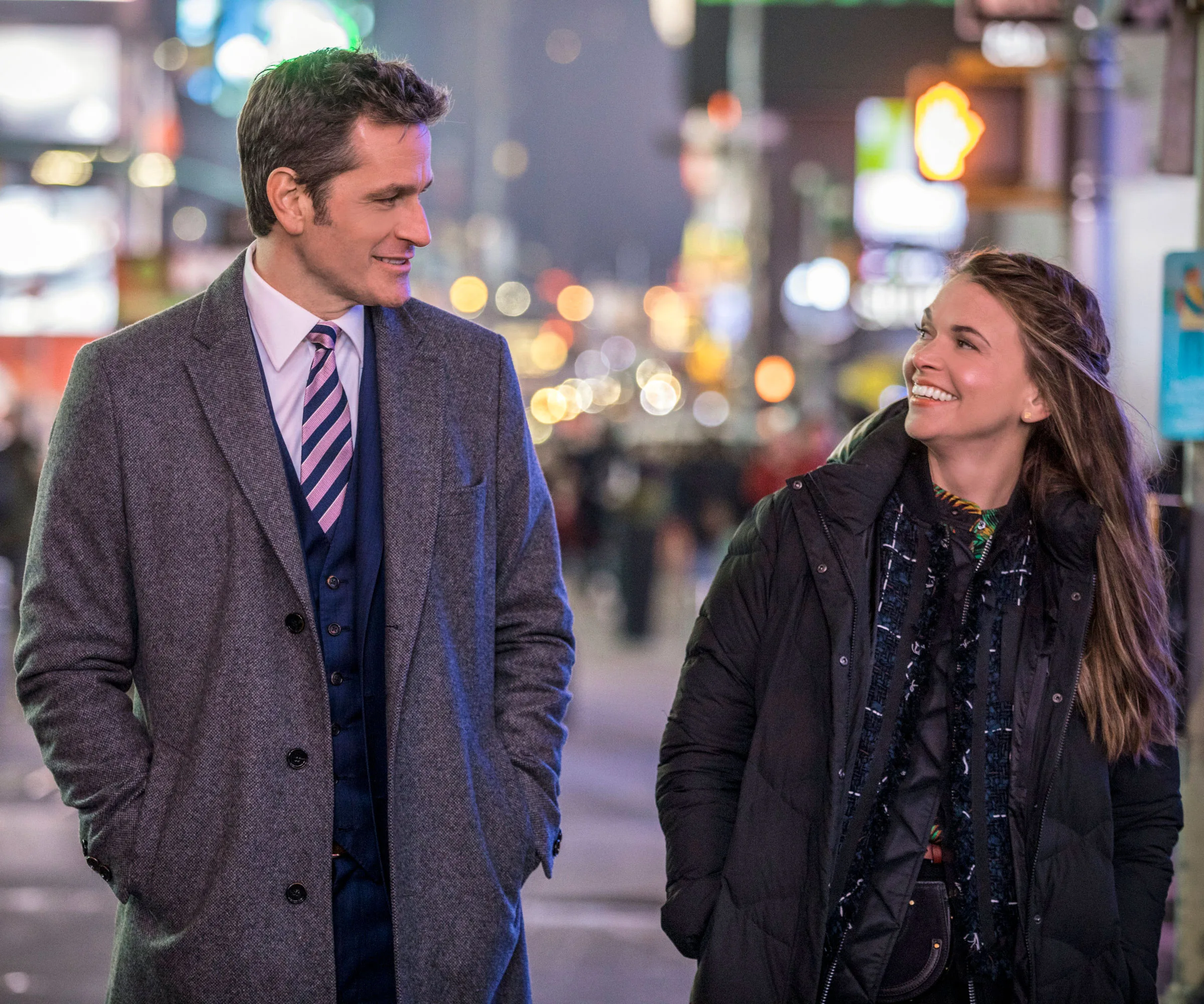 Two people smiling and walking in a city at night, with bright lights blurred in the background.
