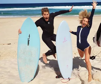Two people in wetsuits joyfully jump on a beach, holding surfboards with ocean waves in the background.