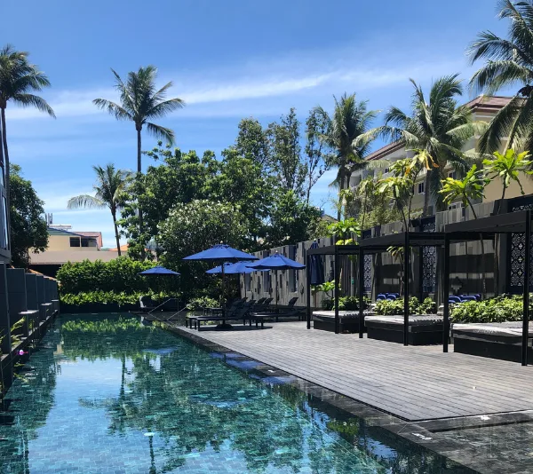 Outdoor swimming pool surrounded by palm trees, sun loungers, and blue umbrellas on a sunny day.