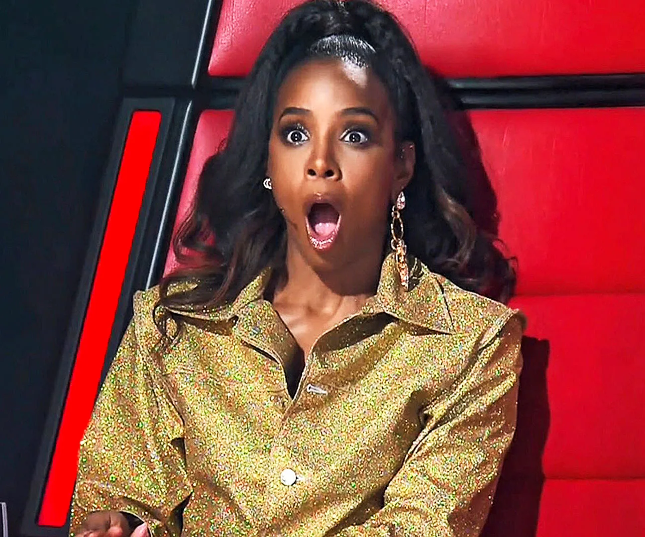Judge on The Voice Australia, surprised in golden outfit, red chair background.