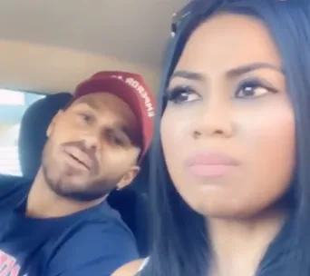 Two people in a car, one in a cap talking, the other looking away with a concerned expression.