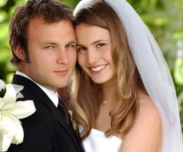 A bride and groom smiling at their wedding, with the bride in a white dress and veil, holding flowers.