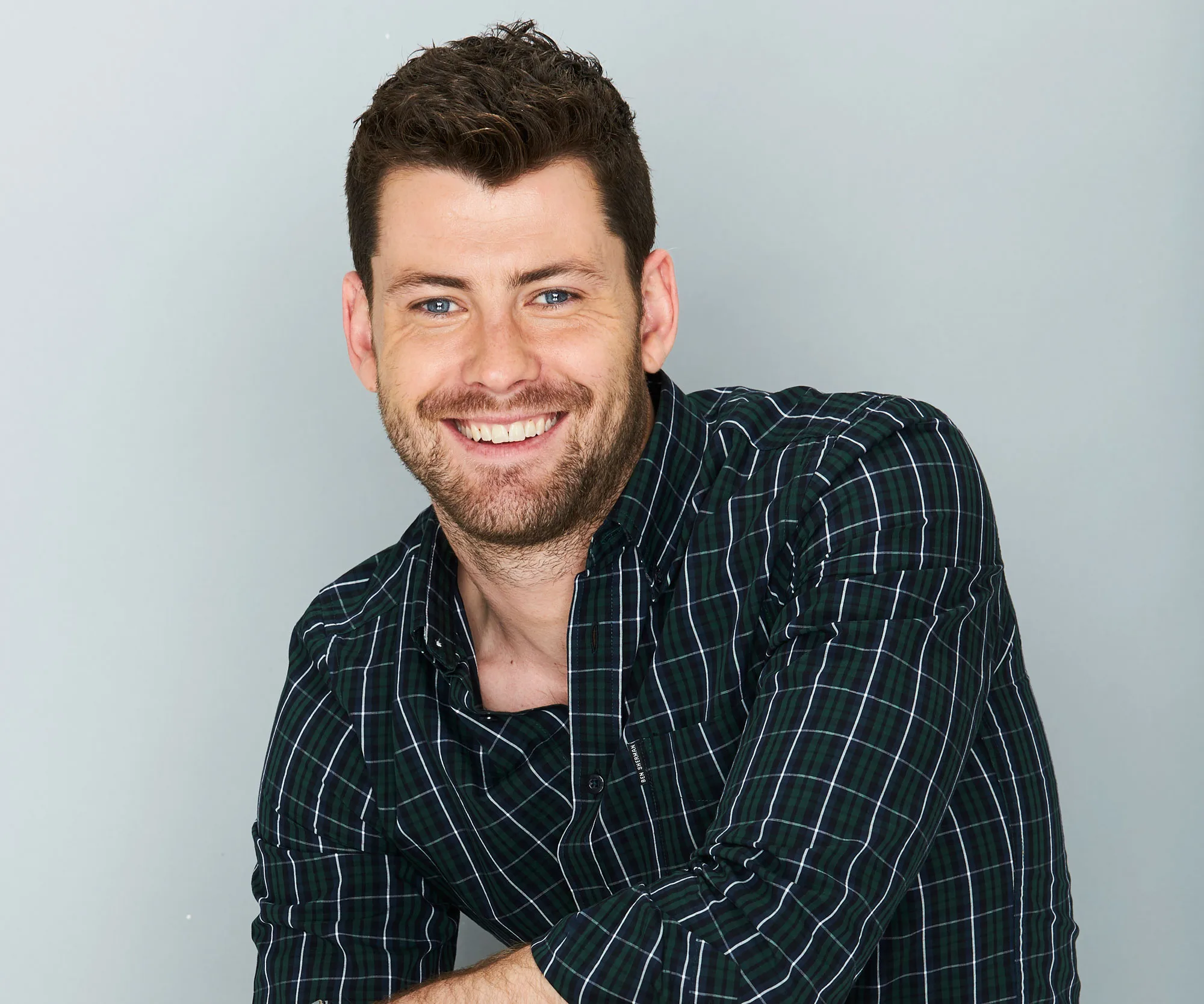 A man with a beard and plaid shirt smiles at the camera, against a plain gray background.